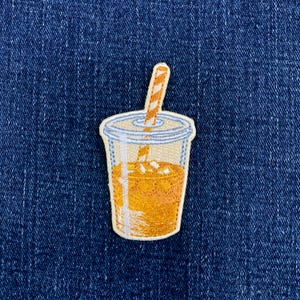 May include: An embroidered patch depicting a clear plastic cup filled with an orange liquid and ice cubes. The cup features a lid and a tall, orange and white striped straw extending upwards, all rendered in detailed thread work on a blue fabric.