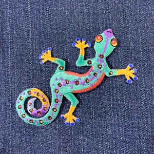 May include: A colorful embroidered gecko patch with green, blue, yellow, and purple accents. The gecko is in a curled position with its tail wrapped around its body.