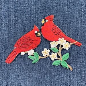 May include: Two red cardinal birds perched on a branch with white flowers. The birds are embroidered on a blue denim background.