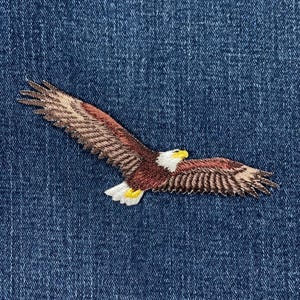 May include: Embroidered patch of a bald eagle in flight, featuring brown, white, and yellow details. The eagle is set against a blue denim background, showcasing the bird's wingspan and detailed feathering.