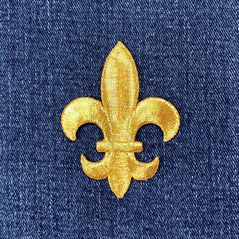 May include: A gold fleur-de-lis patch embroidered on a blue denim background. The patch features a detailed design with a textured appearance, showcasing the classic three-petal symbol. The patch is a decorative element.