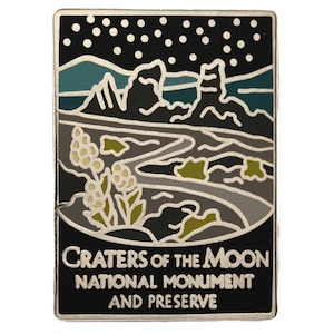May include: A rectangular enamel pin featuring a stylized landscape of the Craters of the Moon National Monument and Preserve. The design includes a night sky with stars, mountains, a winding river, and vegetation. The text "CRATERS OF THE MOON NATIONAL MONUMENT AND PRESERVE" is at the bottom.