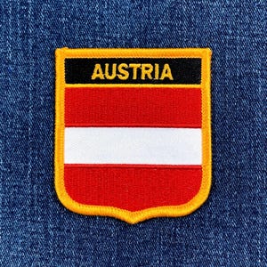 May include: Embroidered patch featuring the Austrian flag design. The patch has a shield shape with a gold border. The flag design includes red and white horizontal stripes, with the word "AUSTRIA" in gold letters on a black banner.