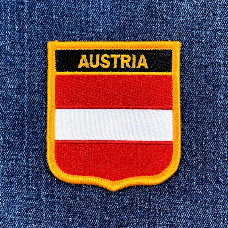 May include: Embroidered patch featuring the Austrian flag design. The patch has a shield shape with a gold border. The flag design includes red and white horizontal stripes, with the word "AUSTRIA" in gold letters on a black banner.