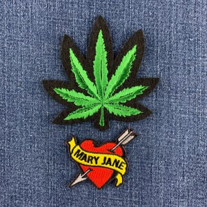 May include: An embroidered patch shaped like a green cannabis leaf with a black border, positioned above a red heart-shaped patch pierced by an arrow, featuring a yellow banner with the black text MARY JANE wrapped around the heart.