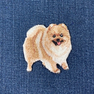 May include: Embroidered patch of a Pomeranian dog in tan and cream hues. The dog has a fluffy coat, a smiling face, and its tongue is visible. The patch is set against a blue denim background.