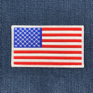 May include: Embroidered patch featuring the American flag. The patch has a white border and is rectangular in shape. The flag design includes a blue field with white stars and red and white stripes.