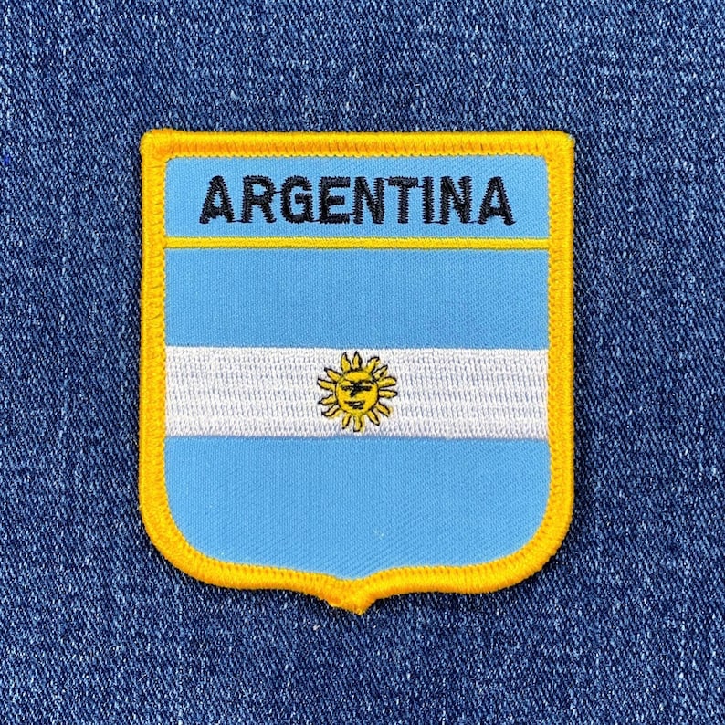 May include: A blue and white embroidered patch with a yellow border. The patch features the Argentinian flag with a sun in the center and the word "ARGENTINA" at the top.