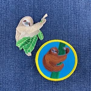 May include: Two embroidered patches featuring sloths. One patch is a beige sloth clinging to a large green leaf. The second patch is a circular design with a yellow border, featuring a brown sloth climbing a green branch against a blue background.