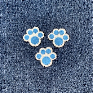 May include: Three embroidered paw print patches. Each patch features a light blue paw print with a white border and a darker blue center, set against a denim blue background.