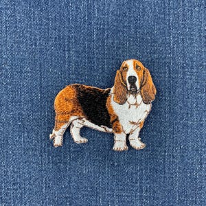 May include: An embroidered patch featuring a Basset Hound dog with a tri-colour coat of black, brown, and white. The dog is depicted in a standing profile position with long ears, a white chest, and a black saddle pattern on its back.