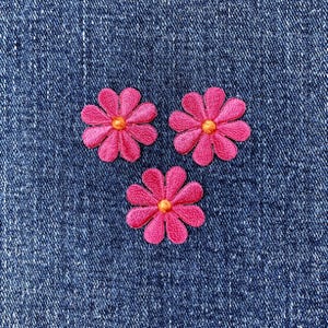May include: Three embroidered pink daisy flower patches with yellow centers are arranged on a blue denim background. The flowers have detailed stitching and are evenly spaced, creating a cheerful, decorative design.