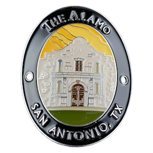 Può includere: Una placca souvenir in metallo di forma ovale con una rappresentazione in smalto in rilievo di The Alamo a San Antonio, Texas. Il design include un edificio beige su un cielo giallo, inserito in un bordo nero con testo argentato THE ALAMO e SAN ANTONIO, TX.