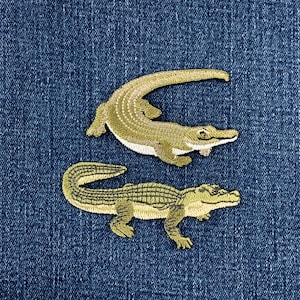 May include: Two embroidered alligator patches in shades of olive green and cream, featuring detailed scale textures and defined limbs. The patches are positioned one above the other against a dark blue denim fabric background with a visible weave.