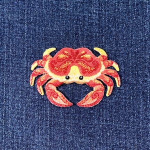 May include: Embroidered crab patch in red, yellow, and orange, with black eyes. The crab patch is on a blue denim background. This patch is suitable for clothing, bags, or other fabric items.
