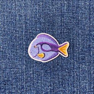 May include: A colorful embroidered patch featuring a stylized fish. The fish has a purple body, a dark purple eye marking, and a yellow tail and accents. The patch is set against a blue denim background.