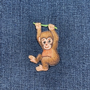 Peut inclure: Écusson brodé représentant un singe brun suspendu à une liane verte. Le singe a un visage brun clair et une poitrine rose, sur un fond en denim bleu. Parfait pour personnaliser des vêtements ou des accessoires.