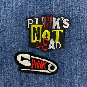 May include: Two embroidered patches. The top patch is black with the text PUNK'S NOT DEAD in white, yellow, and red lettering. The bottom patch is black and shaped like a safety pin with the word PUNK embroidered in red across the center.