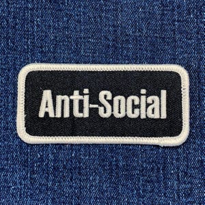 Könnte beinhalten: Schwarz-weißer gestickter Aufnäher mit weißem Rand. Der Aufnäher trägt die Aufschrift "Anti-Social" in weißen Buchstaben.