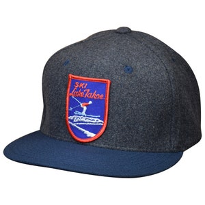 May include: A dark gray wool blend baseball cap with a blue brim. The cap features a red, white, and blue embroidered patch that says "SKI Lake Tahoe" with a skier on a mountain.