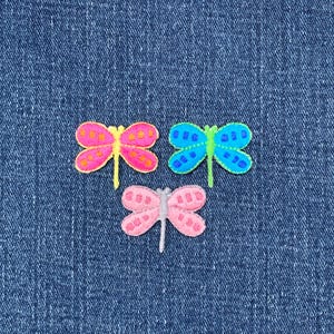 May include: Three embroidered dragonfly patches in pink, blue, and pink with yellow, green, and silver accents. The patches are on a blue denim background. Each dragonfly has detailed wing patterns.