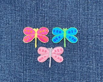 May include: Three embroidered dragonfly patches in pink, blue, and pink with yellow, green, and silver accents. The patches are on a blue denim background. Each dragonfly has detailed wing patterns.