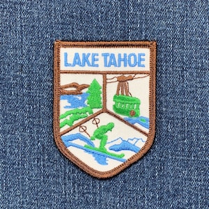 May include: A brown embroidered patch with the text "LAKE TAHOE" in blue and white. The patch features a green pine tree, a blue lake, a white mountain range, and a green cable car.