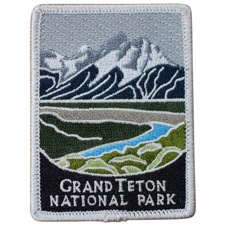May include: Embroidered patch featuring a scenic view of the Grand Teton National Park. The patch depicts a mountain range with snow-capped peaks, a blue lake, and green vegetation. The text "Grand Teton National Park" is embroidered in white on a black background.
