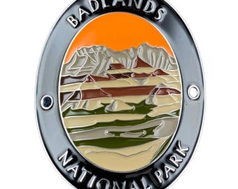 May include: An oval-shaped metal badge featuring a landscape illustration of Badlands National Park. The design includes layered rock formations in shades of tan, brown, and green under a bright orange sky, with the text BADLANDS NATIONAL PARK.