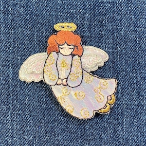 May include: A white angel with gold halo and wings, wearing a white dress with gold accents. The angel has red hair and is holding its hands together in prayer.