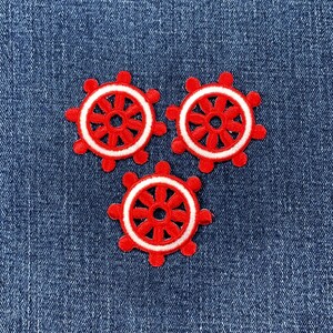 May include: Three red and white ship wheel patches on a blue denim background. Each patch features a red wheel with white detailing. The patches are arranged in a triangular pattern, perfect for nautical-themed crafts.