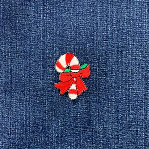 May include: A small embroidered patch featuring a candy cane with red and white diagonal stripes, accented by a large, bright red bow and a small green leaf detail. The patch is designed with a clear black outline and a textured fabric finish.