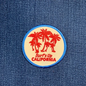 May include: A circular patch with a cream centre, displaying red palm trees and the words "Surf's Up CALIFORNIA". It has a blue border, set against a blue denim background. Suitable for attaching to clothing or accessories.
