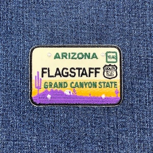 May include: Embroidered patch featuring a design inspired by an Arizona license plate. The patch includes the text "ARIZONA", "FLAGSTAFF", and "GRAND CANYON STATE". The design incorporates purple, yellow, and green colors, with a Route 66 symbol.