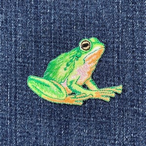 May include: Embroidered frog patch in shades of green, orange, and yellow. The frog has black eyes and is set against a blue denim background. This patch is suitable for clothing or accessories.