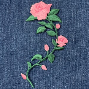 May include: Pink embroidered rose with green leaves and stem on a blue denim background.