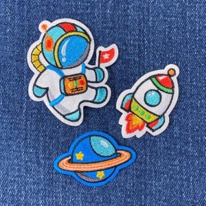 May include: Three embroidered iron-on patches featuring a blue and white astronaut holding a red flag, a white and blue rocket with orange flames, and a blue planet with an orange ring.
