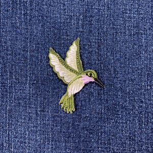 May include: Embroidered hummingbird patch with green, white, and pink details. The patch is on a blue denim background. The hummingbird is depicted in flight, with its wings spread.