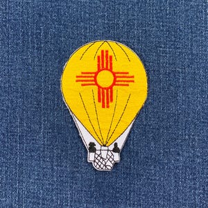 May include: A yellow hot air balloon with a red and white symbol of the state of New Mexico. The balloon is embroidered on a blue denim fabric.