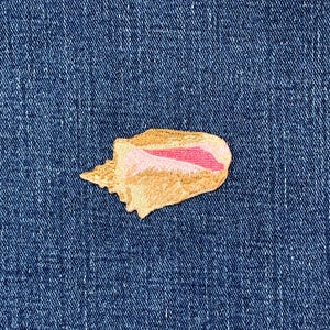 May include: Embroidered seashell patch in tan and pink hues, set on a blue denim backdrop. The patch displays intricate stitching, highlighting the shell's form and texture. Perfect for adding a seaside theme to garments or accessories.