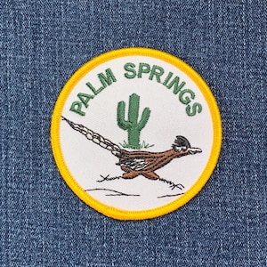 May include: A yellow and white embroidered patch with the text "PALM SPRINGS" in a circle. A cartoon roadrunner with a green cactus in the background is embroidered on the patch.