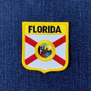 Könnte beinhalten: Gestickter Aufnäher mit dem Design der Flagge des US-Bundesstaates Florida. Der Aufnäher hat einen gelben Rand mit dem Wort "FLORIDA" in Schwarz. Das Flaggen-Design umfasst einen weißen Hintergrund mit roten Diagonalstreifen und einem zentralen Emblem.