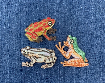 May include: Three embroidered frog patches featuring detailed stitching. One frog is red and yellow, one is white with brown stripes, and one is orange and green. Each patch depicts a frog in a different pose with visible eyes and limbs.