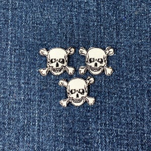 May include: Three white skull and crossbones patches on a blue denim background. Each patch features a detailed skull with crossbones beneath. The patches are evenly spaced, creating a repeating pattern.