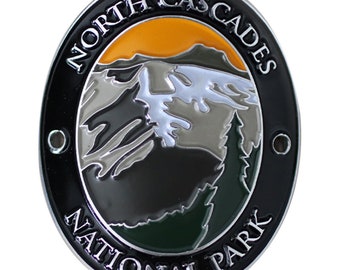 May include: A silver oval-shaped metal pin with a black background. The pin features a colorful illustration of a mountain range with a sunset in the background. The text "NORTH CASCADES NATIONAL PARK" is written around the edge of the pin.