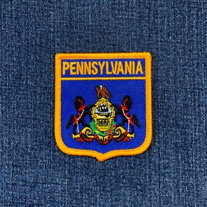 May include: A Pennsylvania state patch with a blue background and a gold border. The word "PENNSYLVANIA" is in gold letters at the top. The state seal is in the center. The patch is on a blue denim background.