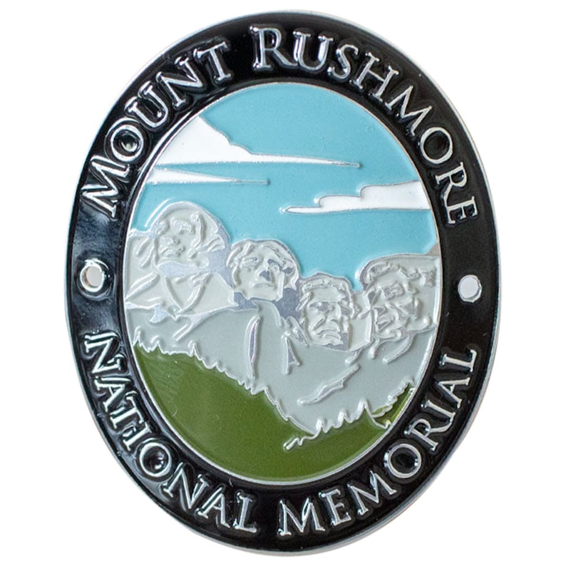 May include: A black and silver metal pin with a green, white, and blue image of Mount Rushmore National Memorial. The text "MOUNT RUSHMORE NATIONAL MEMORIAL" is written around the edge of the pin.