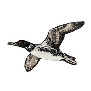 May include: An embroidered patch depicting a bird in flight with black, white, and grey thread detailing. The bird features a distinct red eye, a pointed beak, and wings spread wide, showcasing intricate stitching patterns across its body.