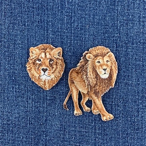 May include: Two embroidered patches featuring lion designs. One patch shows a detailed lion head facing forward with golden brown fur. The second patch depicts a full-body lion in a walking stance, showcasing a thick mane and textured fur details.
