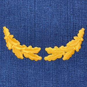 May include: Two embroidered golden-yellow leaf branch patches arranged in a semi-circle on a blue denim background. The patches are detailed with veining and have a textured appearance, suitable for sewing or ironing onto fabric.
